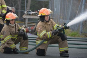 Two firefighters kneeling down one behind the other working together to support a fire hose. 