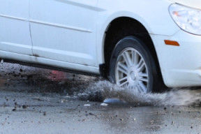 A white car drives through a pothole filled with water, splashing as it passes.