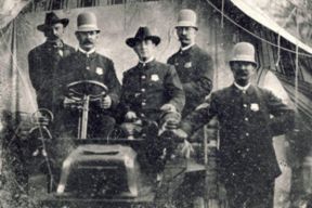Black and white historic posed photo of a uniformed officer sitting in a car with four uniformed officers standing beside the car.
