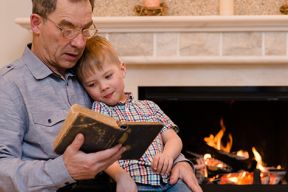 Image of a Grandfather reading a book to his Grandson sitting on his lap in front of a fire.