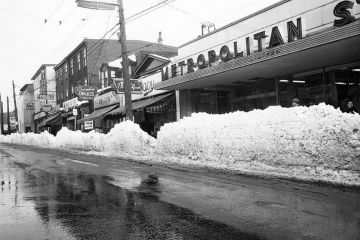 Black and white image of the east side of Gottingen Street, after a heavy snowfall in February 1960.  Shows Metropolitan Stores, J. Macy Co. Ltd. ladies wear, Coombes Drug Store and Luncheonette, Withrows Drugs, and [Allen and Son Ltd. hardware