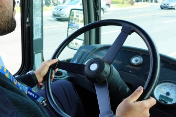 A bus Operator drives a bus, with the wheel of the bus visible over the Operator's shoulder.
