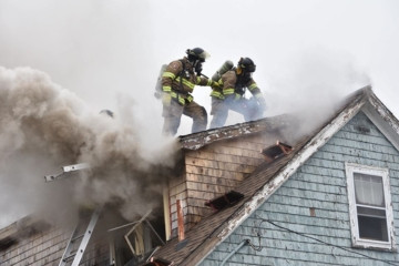 Two firefighters on a roof top of a house that is on fire. Smoke is visibly coming out of the roof.